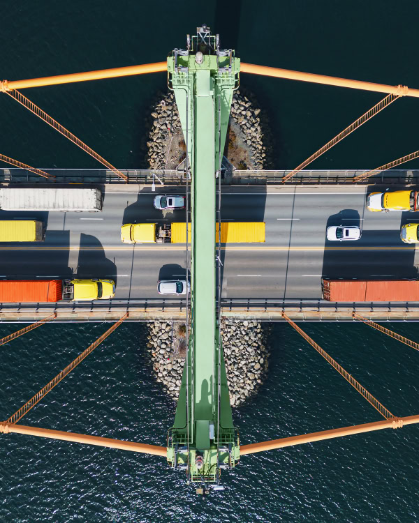 Aerial view of vehicles, including trucks and cars, crossing a suspension bridge over water, with orange cables and a central green support structure.