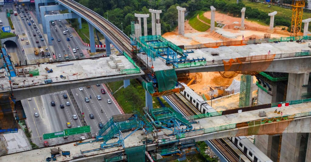 Aerial view of a highway next to a train track under construction with multiple elevated concrete structures and scaffolding. Vehicles are visible on the highway, surrounded by greenery.