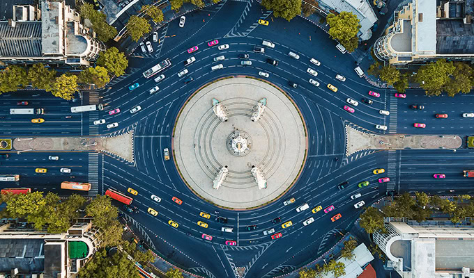 Aerial view of a large roundabout with multiple lanes of cars and buses surrounding a central monument, bordered by trees and nearby buildings.