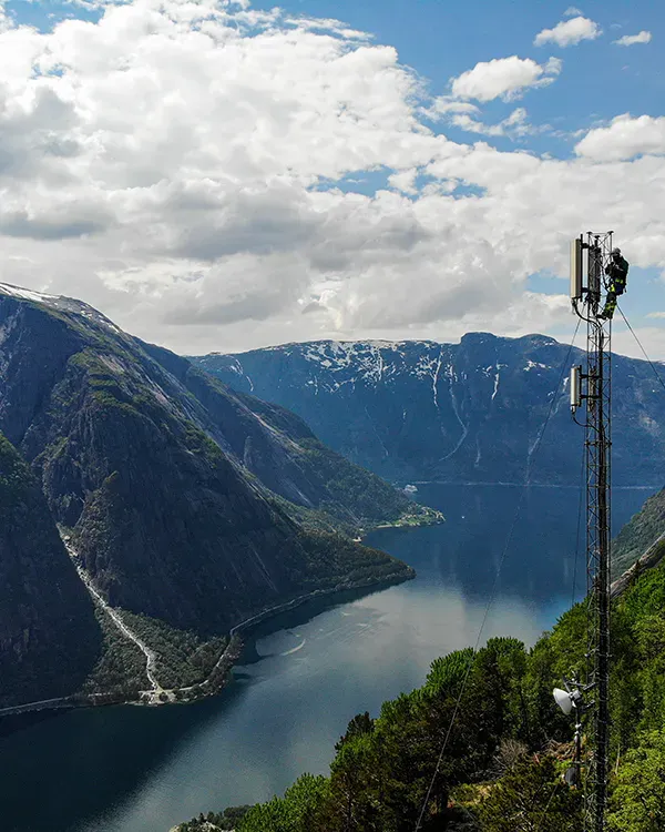 A worker climbs a tall communication tower overlooking a fjord with mountains, winding roads, and a partially cloudy sky in the background.