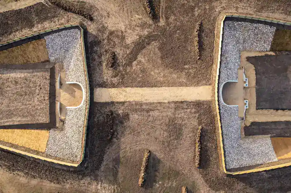Aerial view of two rectangular, gravel-covered bunkers or structures facing each other across a dirt path, set in a dry, brown landscape.